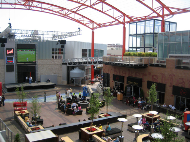 An entertainment district that features restaurants with outdoor seating, a stage, and covered central seating area.
